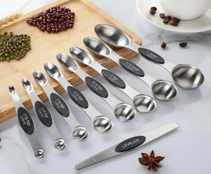 Cuillères doseuses magnétiques en acier inoxydable et araseur disposés sur un plan de travail avec une planche en bois et des haricots